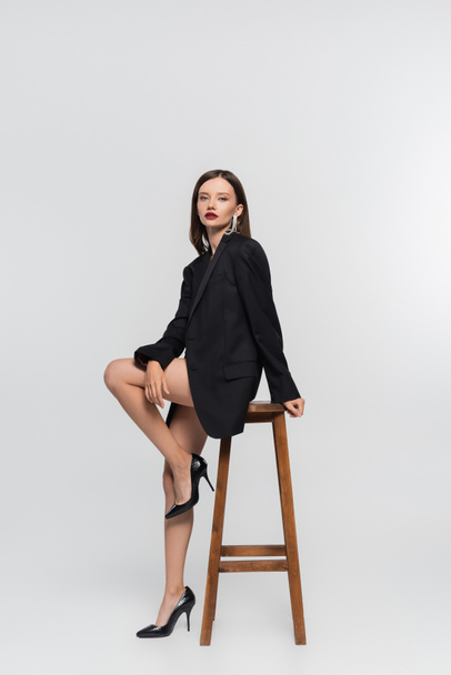 sexy woman in black blazer, with naked legs, looking at camera while sitting on high stool on grey - Fotografie, Obrázek
