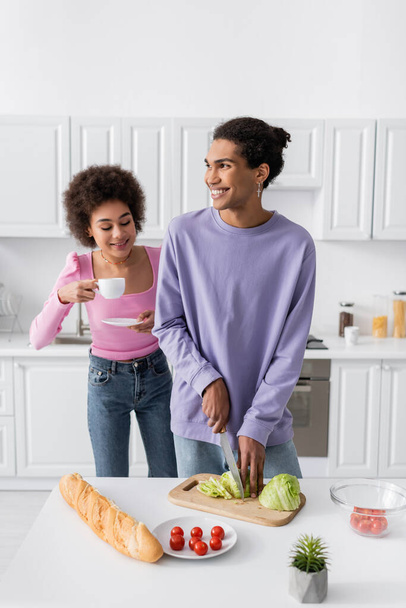 Cheerful african american woman holding cup near boyfriend cutting salad in kitchen  - 写真・画像