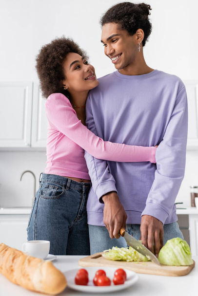 Smiling african american woman hugging boyfriend cutting cabbage in kitchen  - 写真・画像