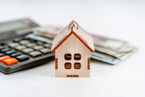 A wooden model of the house stands with a calculator and dollar bills on a white background. Business concept, real estate purchase - 写真・画像