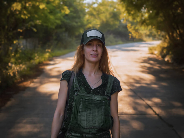 Una mujer con gorra camina por un camino sombreado con un bosque a ambos lados en un día de verano. - Foto, imagen