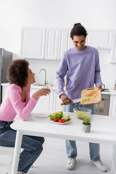 Cheerful man cooking salad near vegetables and girlfriend with coffee in kitchen  - Foto, Bild