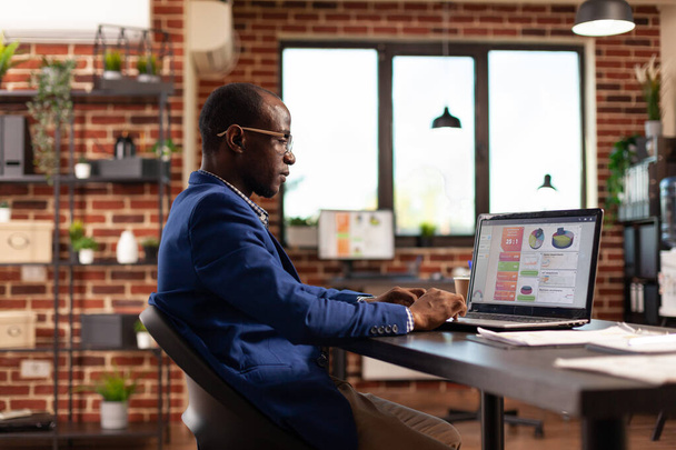 Afro-Amerikaanse werknemer met behulp van laptop om zakelijke grafieken en bedrijfsgroei te analyseren - Foto, afbeelding