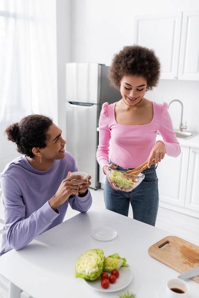 Young african american woman holding bowl with salad near boyfriend with coffee in kitchen  - Foto, Imagen