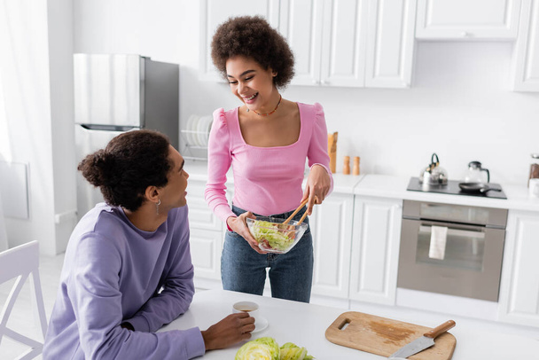 Positive african american woman holding bowl with salad near boyfriend with cup of coffee at home  - Fotoğraf, Görsel