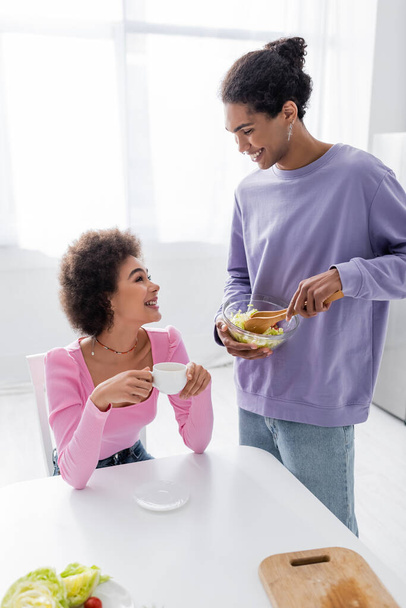 Cheerful african american man holding bowl with salad near girlfriend with coffee at home  - Foto, Bild