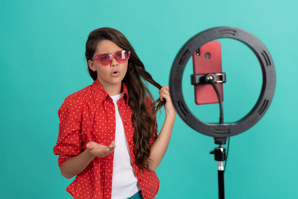 bambino serio parlando sulla macchina fotografica mostra i suoi capelli lunghi blogging online con selfie led, infanzia. - Foto, immagini