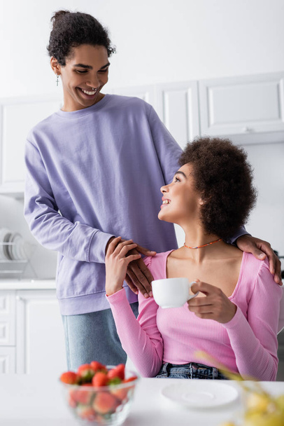 African american man hugging girlfriend with cup near blurred strawberries in kitchen  - Zdjęcie, obraz