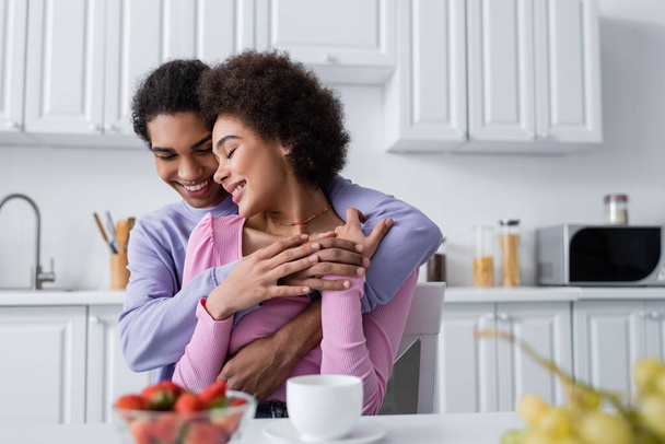 Happy african american man embracing girlfriend near fruits and blurred coffee in kitchen  - Foto, Imagem
