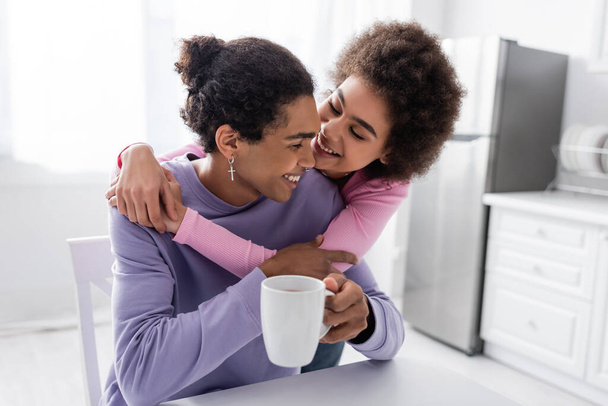 Smiling african american woman hugging boyfriend with cup in kitchen  - Фото, изображение