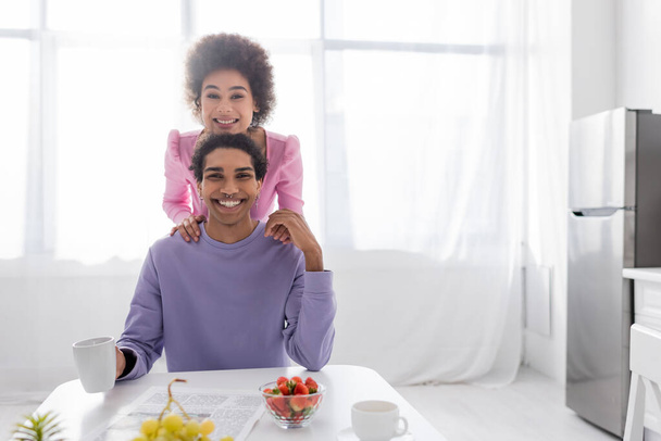 Couple afro-américain positif regardant la caméra près du journal, du café et des fruits dans la cuisine  - Photo, image
