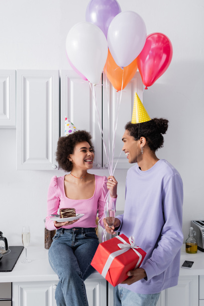 Smiling african american couple with balloons, gift and champagne in kitchen  - Valokuva, kuva