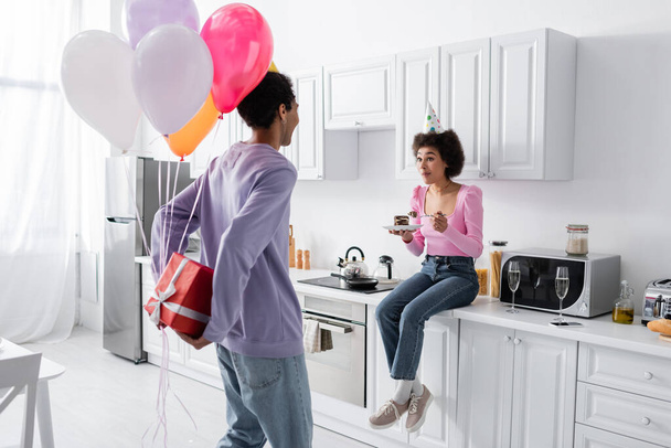 Mladý africký Američan muž skrývá dárek a balónky v blízkosti ohromený přítelkyně s dort doma  - Fotografie, Obrázek