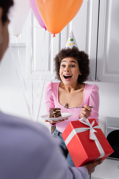 Excited african american woman holding cake near blurred boyfriend with gift and balloons at home  - Фото, изображение