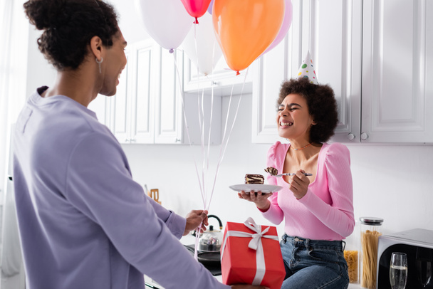 Ευτυχισμένη Αφροαμερικανή γυναίκα κρατώντας τούρτα κοντά στο φίλο της με μπαλόνια και κουτί δώρου στην κουζίνα  - Φωτογραφία, εικόνα