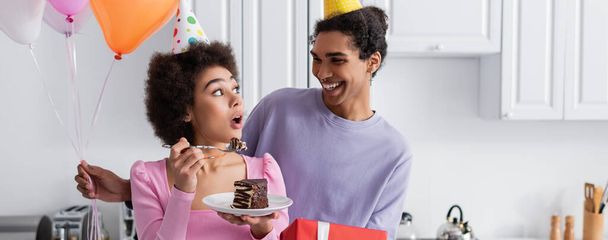 Excited african american woman holding birthday cake near boyfriend with present and balloons at home, banner  - Фото, зображення
