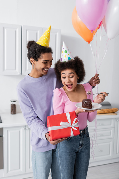 笑顔アフリカ系アメリカ人男持っていますプレゼント近くに驚いていますガールフレンドでパーティーキャップとともに誕生日ケーキ-家庭で  - 写真・画像