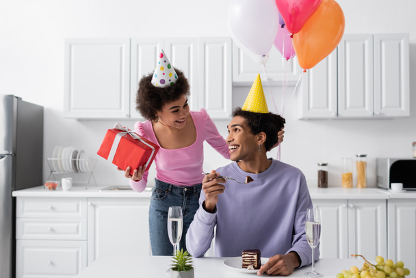 Cheerful african american woman holding present near boyfriend in party cap and birthday cake at home  - Foto, imagen