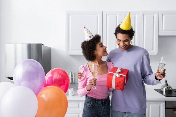 Smiling african american woman in party cap holding gift and champagne near boyfriend and balloons in kitchen  - Foto, immagini