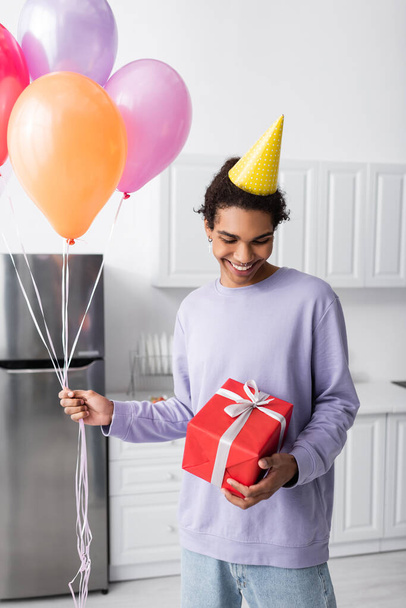Smiling african american man holding present and balloons during birthday party at home  - Фото, изображение