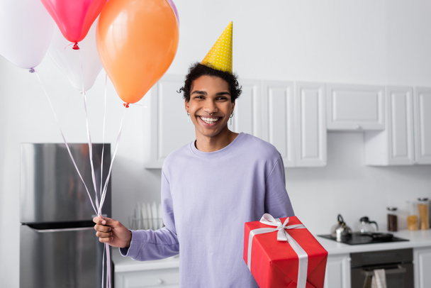 Happy african american man in party cap holding gift and balloons while looking at camera at home  - Photo, Image