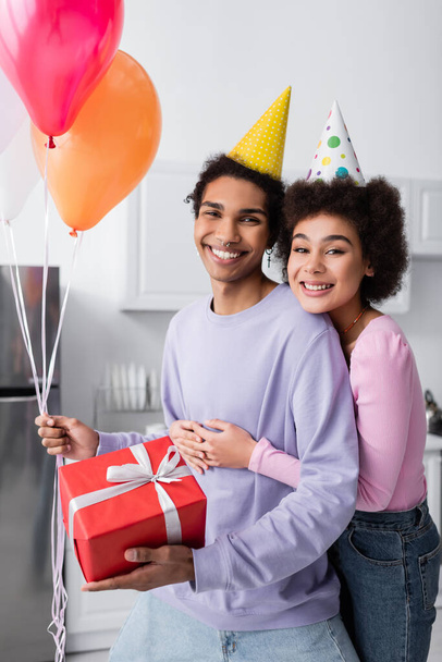 Cheerful african american woman in party cap hugging boyfriend with balloons and present at home  - Фото, изображение