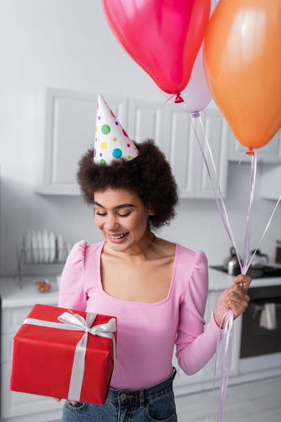 Young african american woman in party cap holding gift and balloons at home  - Photo, image