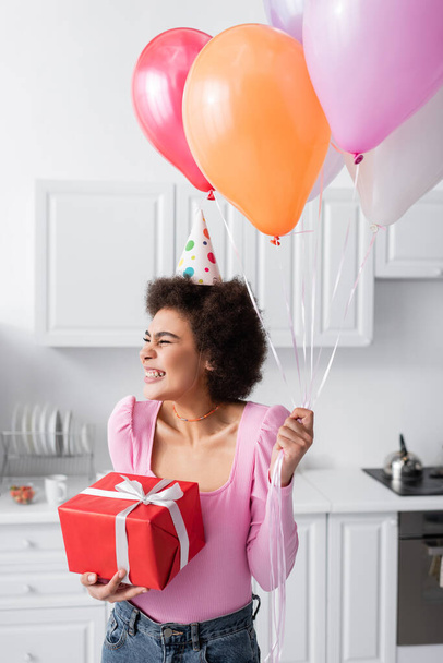Happy african american woman in party cap holding gift and balloons in kitchen at home  - Valokuva, kuva