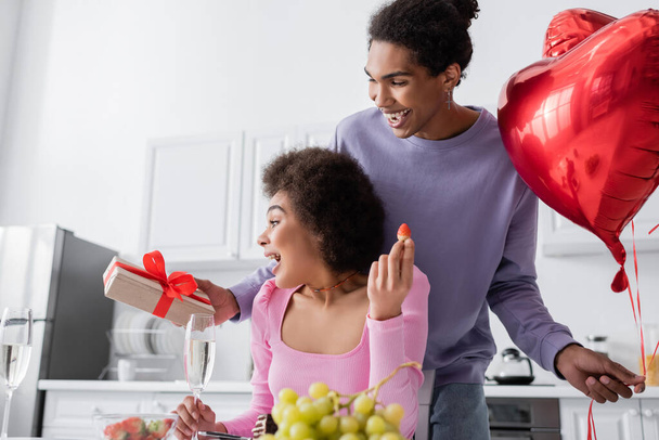 Happy african american man holding heart-shaped balloons and gift near girlfriend with present and strawberry at home  - 写真・画像