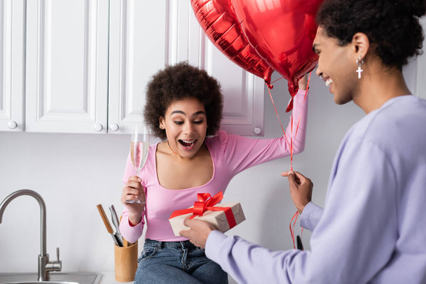 Усміхнений афроамериканець, що тримає повітряні кулі і дарунок біля збудженої подруги з шампанським на кухні.  - Фото, зображення