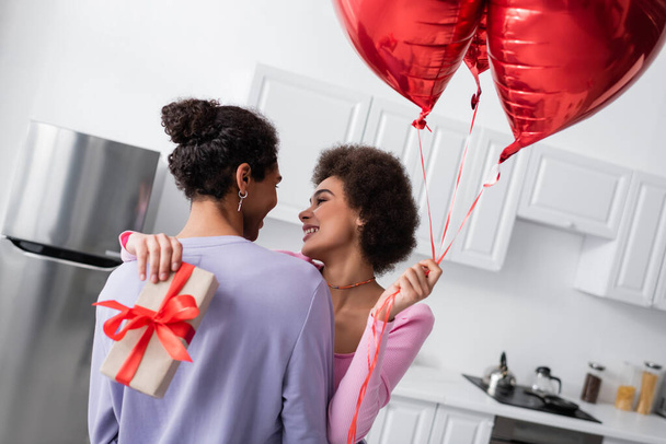 Smiling african american woman holding balloons and present while embracing boyfriend at home  - Fotó, kép
