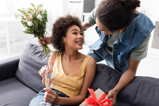 African american man holding bouquet and present near smiling girlfriend with champagne at home  - Foto, afbeelding