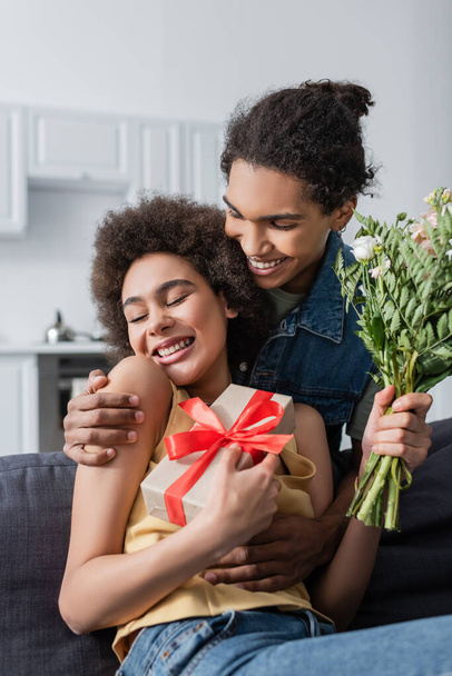 Young african american man hugging girlfriend with flowers and present at home  - Zdjęcie, obraz