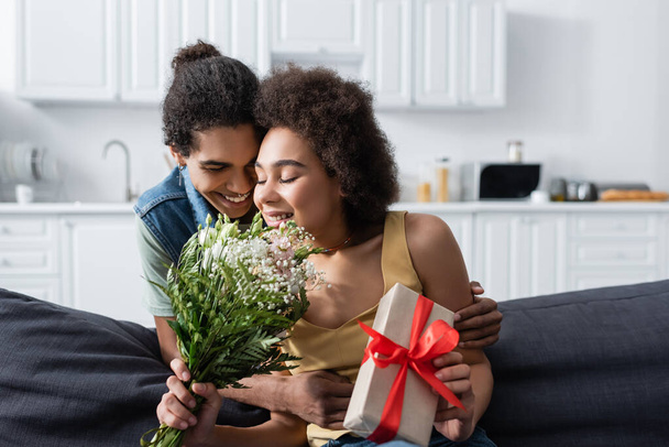 Young african american man hugging smiling girlfriend with bouquet and present at home  - Foto, Imagem
