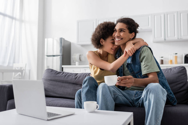 Young african american woman with closed eyes hugging smiling boyfriend with cup near laptop at home  - Foto, Imagem