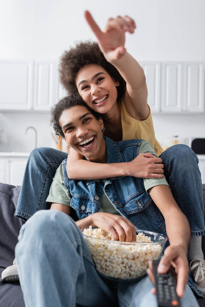 African american woman pointing with finger and hugging boyfriend with remote controller and popcorn at home  - Foto, Imagen