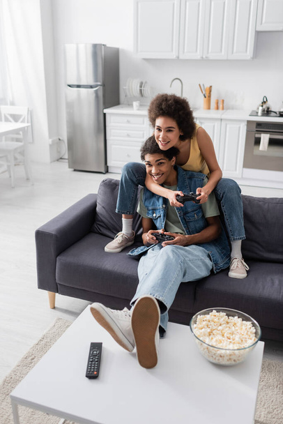 KYIV, UKRAINE - NOVEMBER 9, 2021: Young african american couple playing video game near popcorn and remote controller at home  - Photo, image