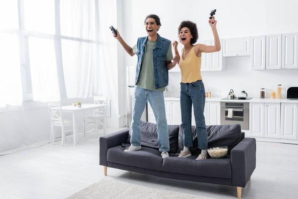KYIV, UKRAINE - NOVEMBER 9, 2021: Excited african american couple with joysticks standing on couch near popcorn and remote controller   - Photo, Image