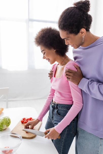 Side view of young african american man hugging girlfriend cutting salad at home  - Foto, Bild