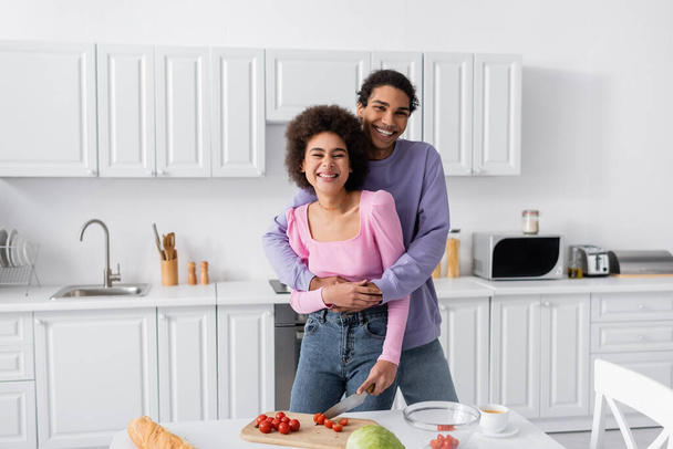 Smiling african american man hugging girlfriend near vegetables and coffee at home  - Foto, Imagen