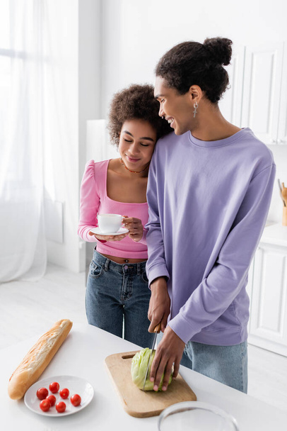 Smiling african american man cutting cabbage near girlfriend with coffee in kitchen  - Photo, Image