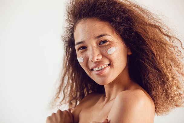 Portrait of woman with lush curly hair and freckles using moisturizing face cream. - Photo, image
