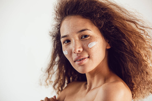 Portrait of woman with lush curly hair and freckles using moisturizing face cream. - Foto, afbeelding