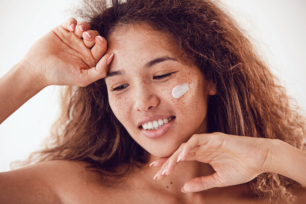 Portrait of smiling woman with lush curly hair and freckles using moisturizing face cream. - Foto, Bild
