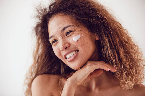 Portrait of smiling woman with lush curly hair and freckles using moisturizing face cream. - Foto, Bild