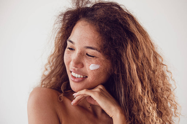 Portrait of smiling woman with lush curly hair and freckles using moisturizing face cream. - Foto, Imagem