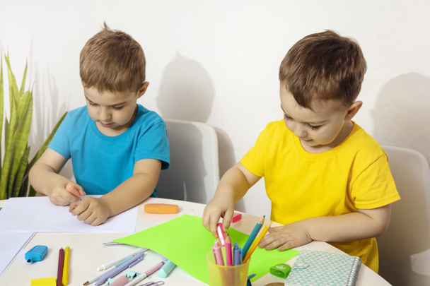 Happy kids with yellow and blue shirts doing arts and crafts together at their desk. Children draw on colored paper. Boys play at home. - Фото, изображение