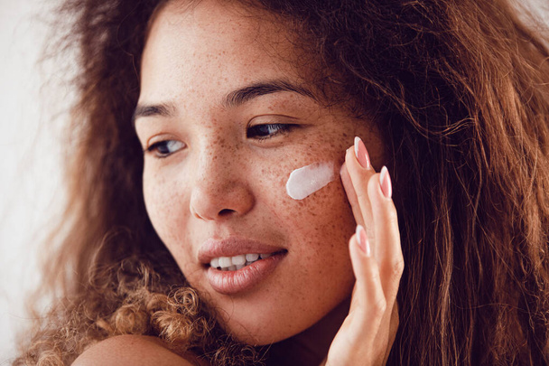 Portrait of woman with curly hair and freckles applying moisturizer to her face. - Fotoğraf, Görsel