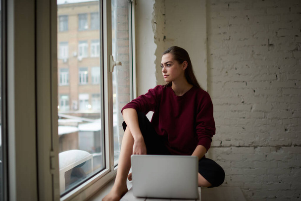 Young concentrated female freelancer in casual outfit with long hair looking away sitting on windowsill in comfortable pose with laptop while doing work - Foto, Imagem