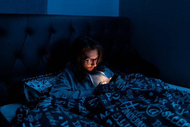 Młody człowiek uzależniony od mediów społecznościowych sprawdza swój telefon przed pójściem spać, Uzależnienie, social media, po ciemnej koncepcji - Zdjęcie, obraz